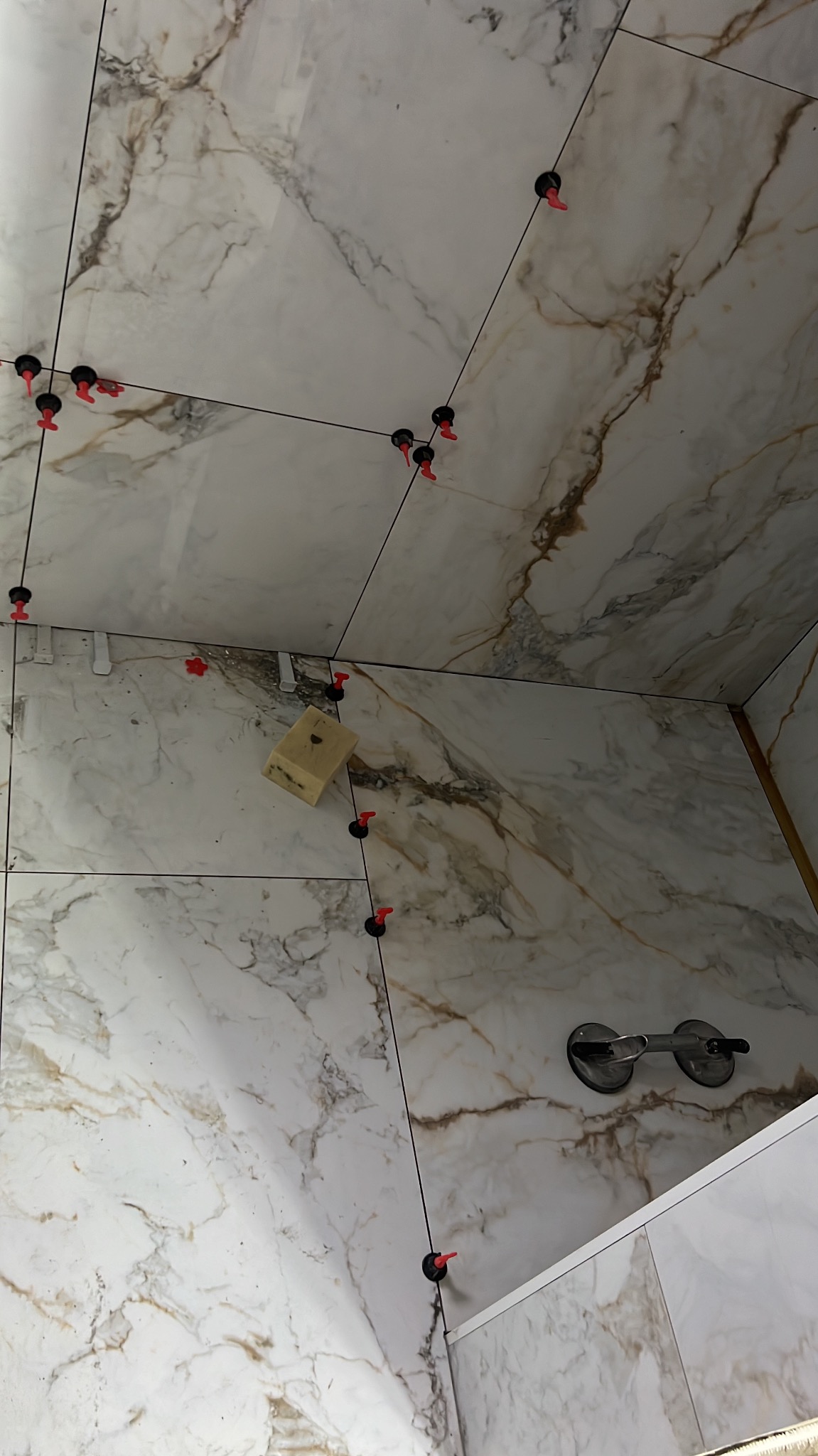 Montaż płytek imitujących marmur na ścianie, widoczne przyssawki do płytek, system poziomowania z czarnymi klipsami i czerwonymi klinami, gąbka do czyszczenia fug.