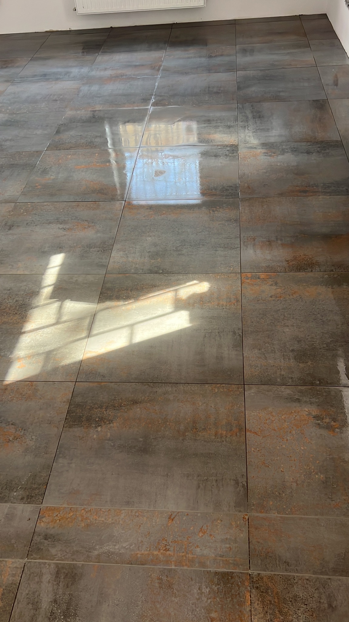Ułożone szare, matowe płytki podłogowe z rdzawymi akcentami, odbijające światło słoneczne wpadające przez okno.