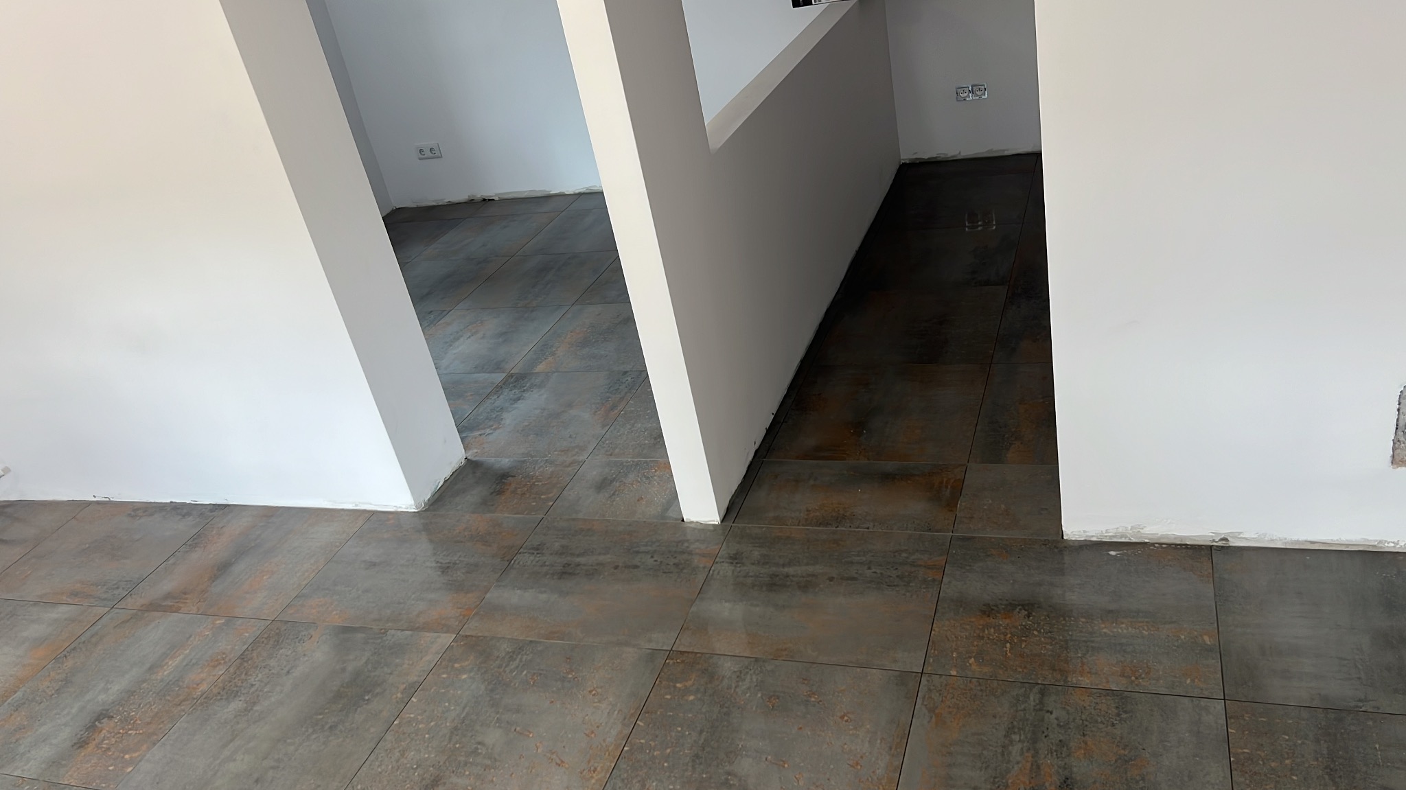 Ułożona podłoga z płytek imitujących metal w odcieniach szarości i brązu, widoczne białe ściany i narożniki, świeżo położone płytki podłogowe, widoczne gniazdka elektryczne.