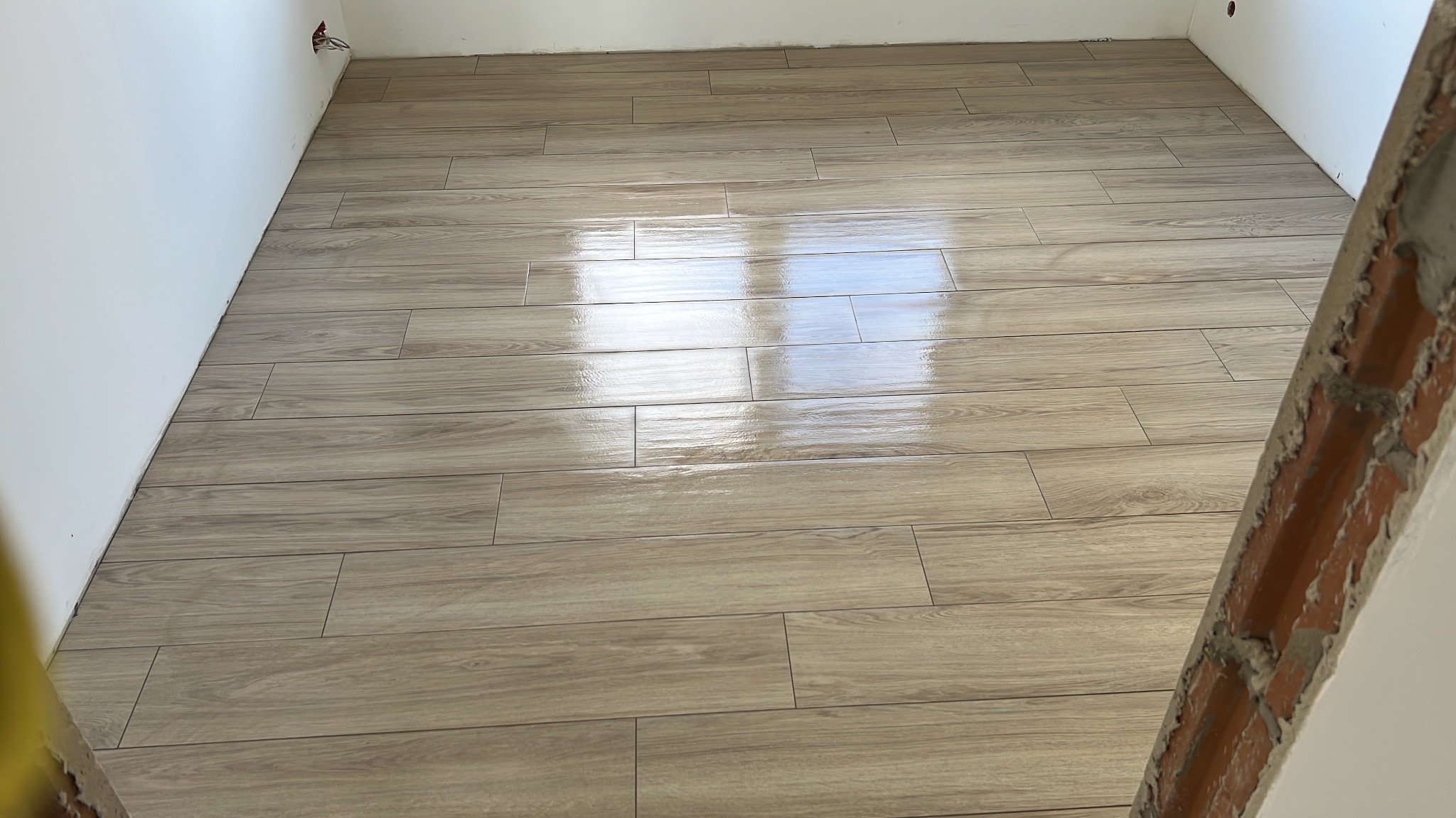 Ułożona jasna podłoga z płytek imitujących drewno, widoczne odbicie światła, fragment surowej ściany z cegły w rogu.