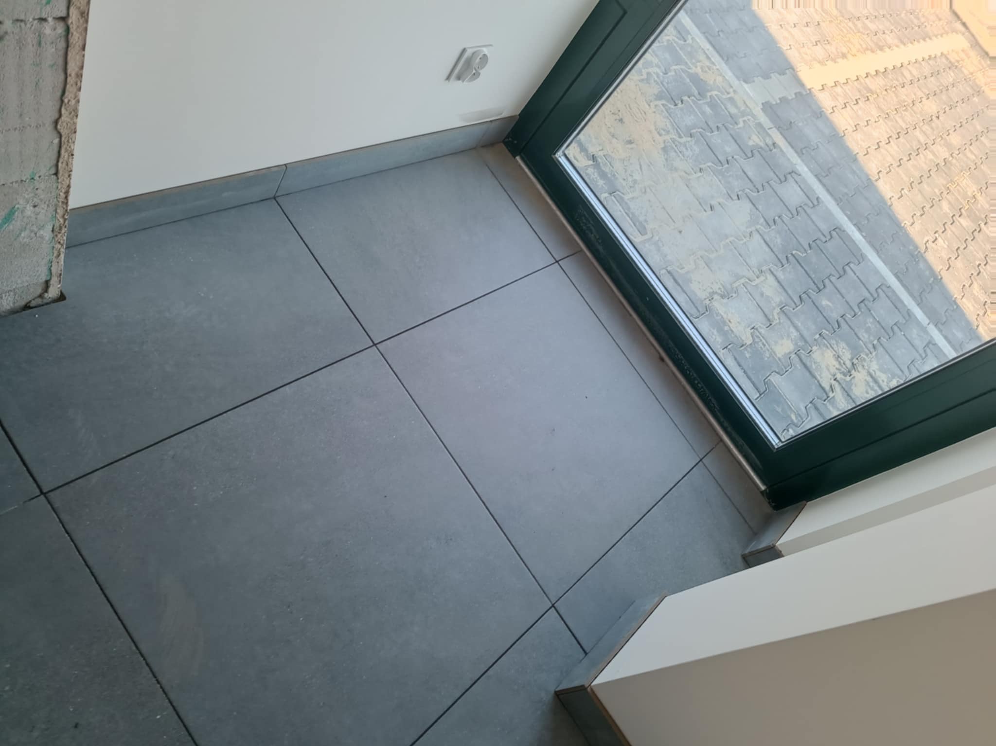 Szare, matowe płytki podłogowe ułożone w regularny wzór, widoczne przy oknie. Fragment ściany z gniazdkiem i surowy fragment ściany w tle. Minimalistyczny design.