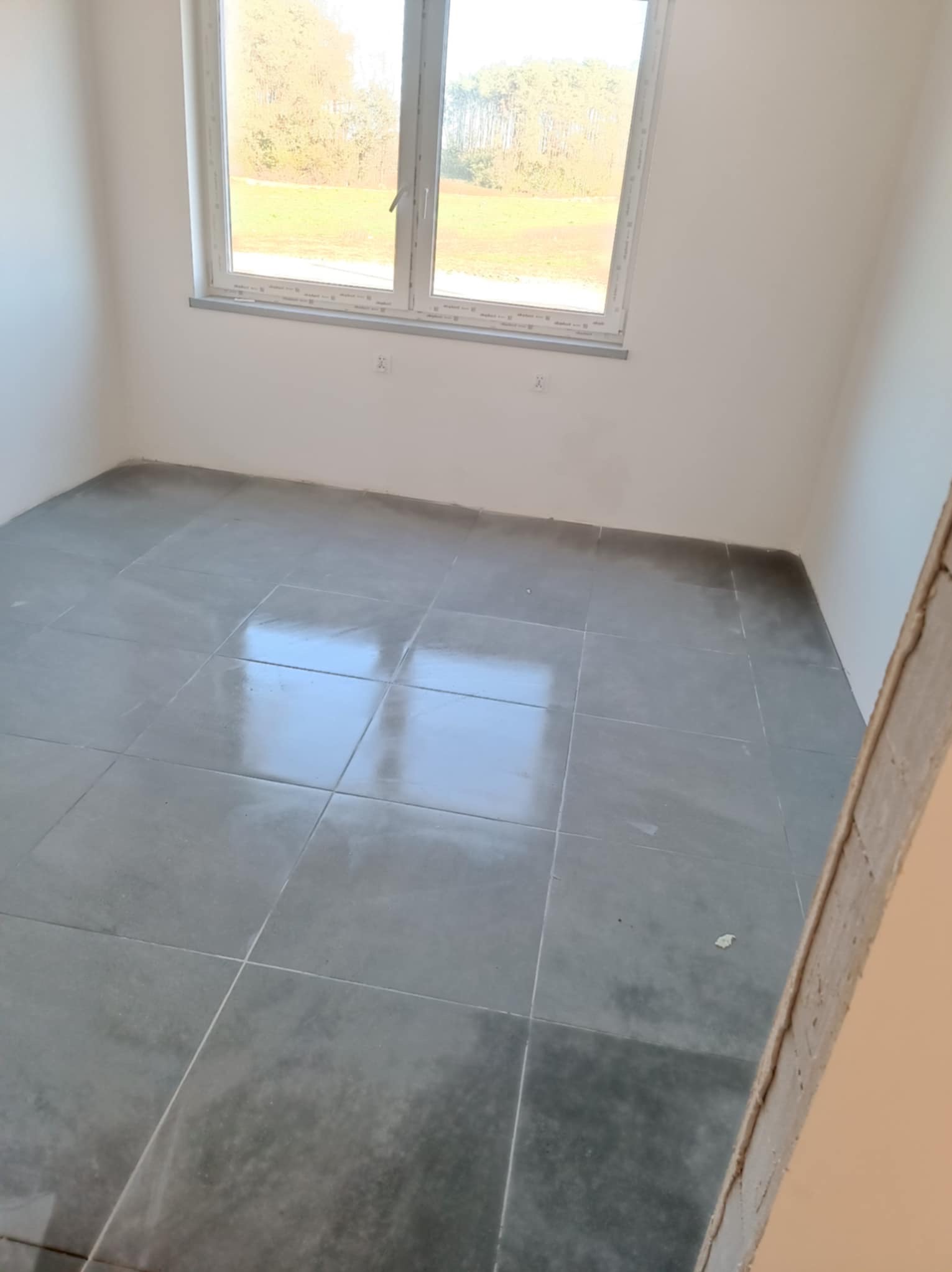 Szare płytki podłogowe w jasnym pokoju z oknem. Widok na zewnątrz przez okno. Świeżo położona podłoga, widoczne fugi. Częściowo widoczna ościeżnica drzwi.