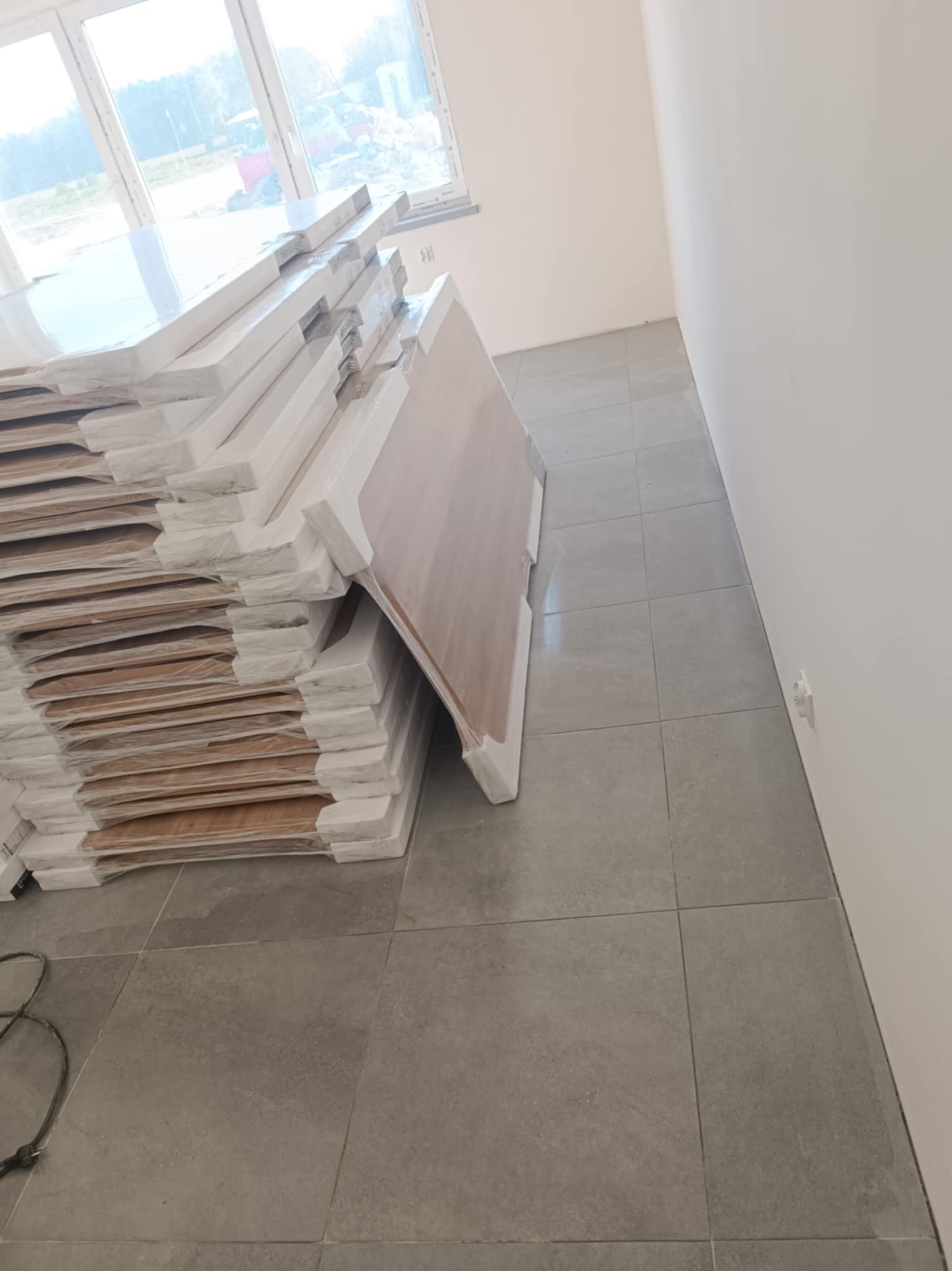 Stos płytek podłogowych w folii ochronnej, ułożonych na szarych płytkach w nowym pomieszczeniu z oknem. Przygotowanie do prac wykończeniowych.