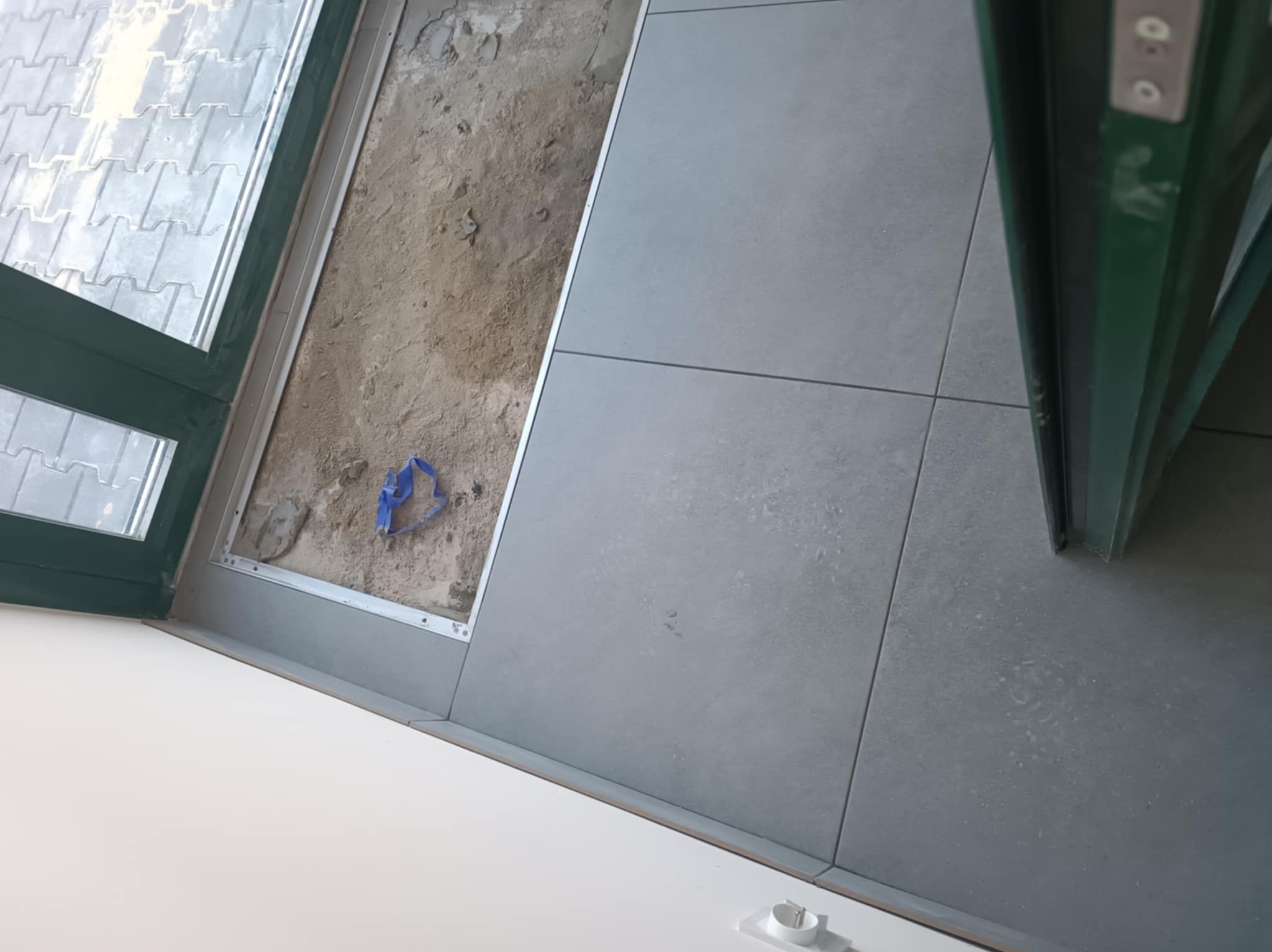 Szare płytki podłogowe ułożone przy oknie, widoczny fragment podłoża przed położeniem płytek. Etap prac wykończeniowych, widok z góry.