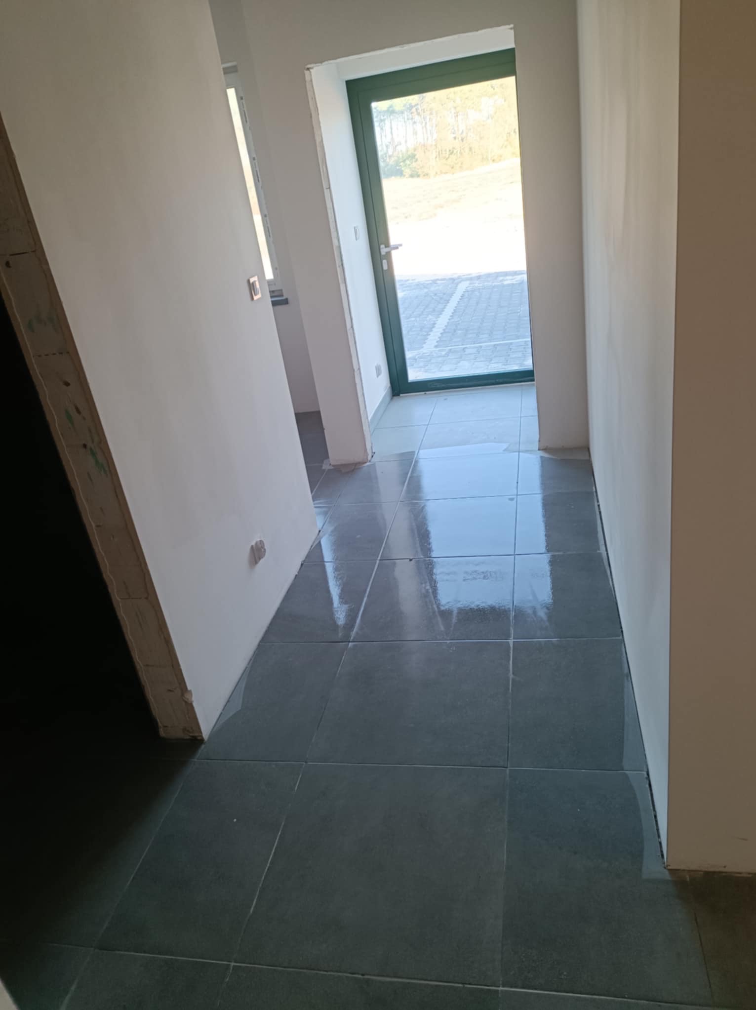 Szare płytki podłogowe w korytarzu z widokiem na zewnątrz przez zielone drzwi. Ściany pomalowane na biało, widoczne niedoskonałości wykończenia.