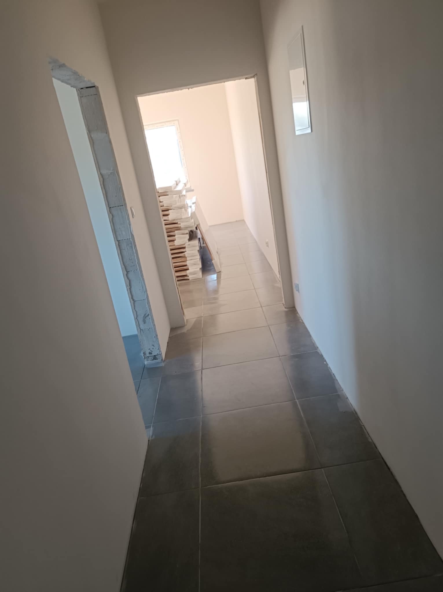 Wykończony korytarz z szarymi płytkami podłogowymi. Widoczne przygotowane do montażu ościeżnice i sterta materiałów budowlanych w głębi. Świeżo pomalowane ściany.