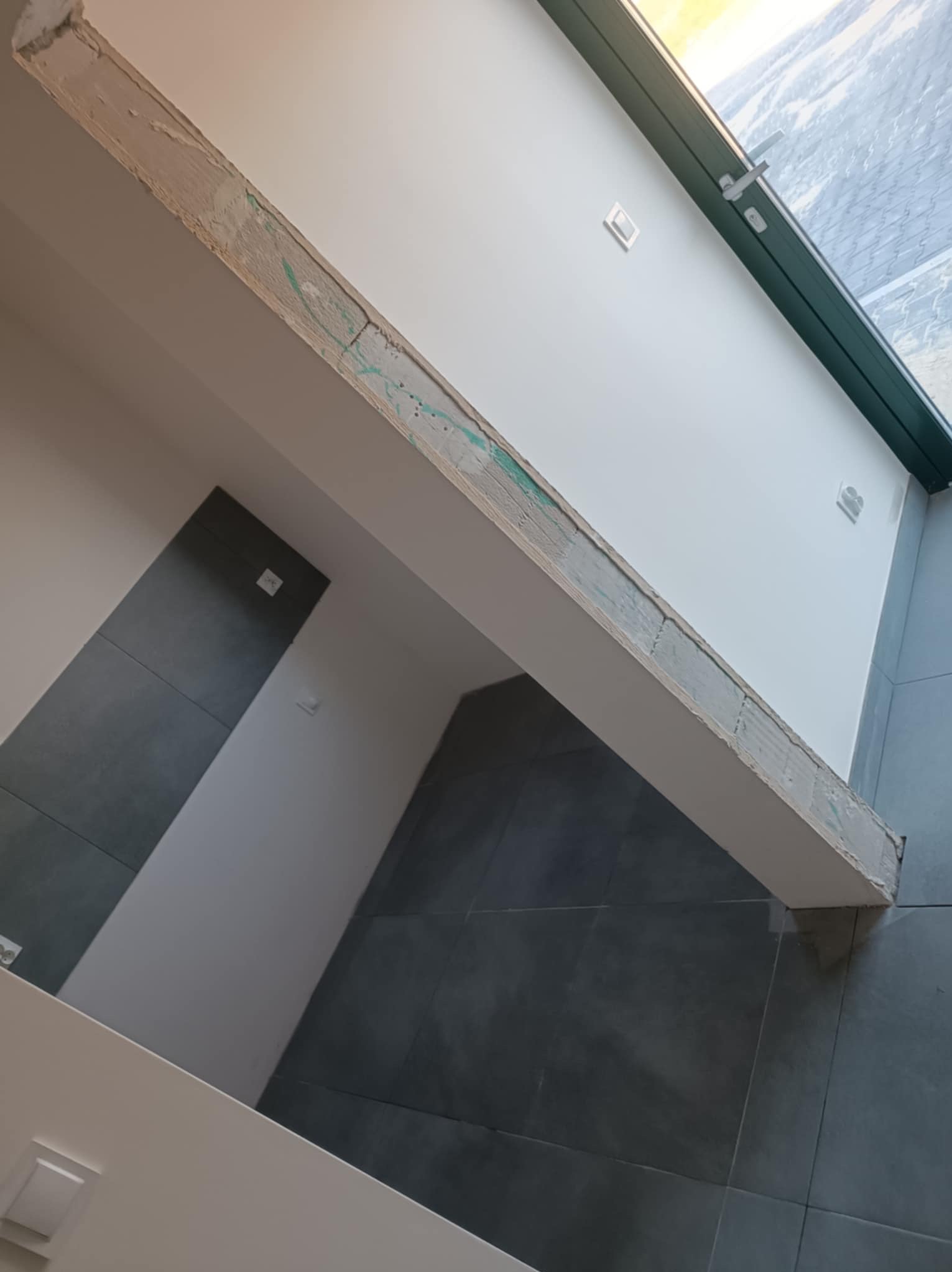 Wnętrze w trakcie wykańczania: szara płytka na ścianie, biała ściana z oknem i belka z nałożoną gładzią, widoczne ślady po szpachlowaniu.