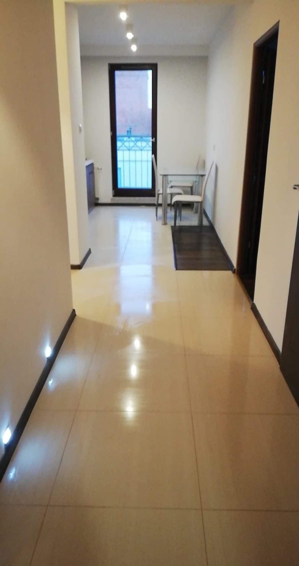 Jasne, błyszczące płytki podłogowe w korytarzu z oświetleniem punktowym przy podłodze. W tle widoczne okno balkonowe i minimalistyczny zestaw stół z krzesłami.