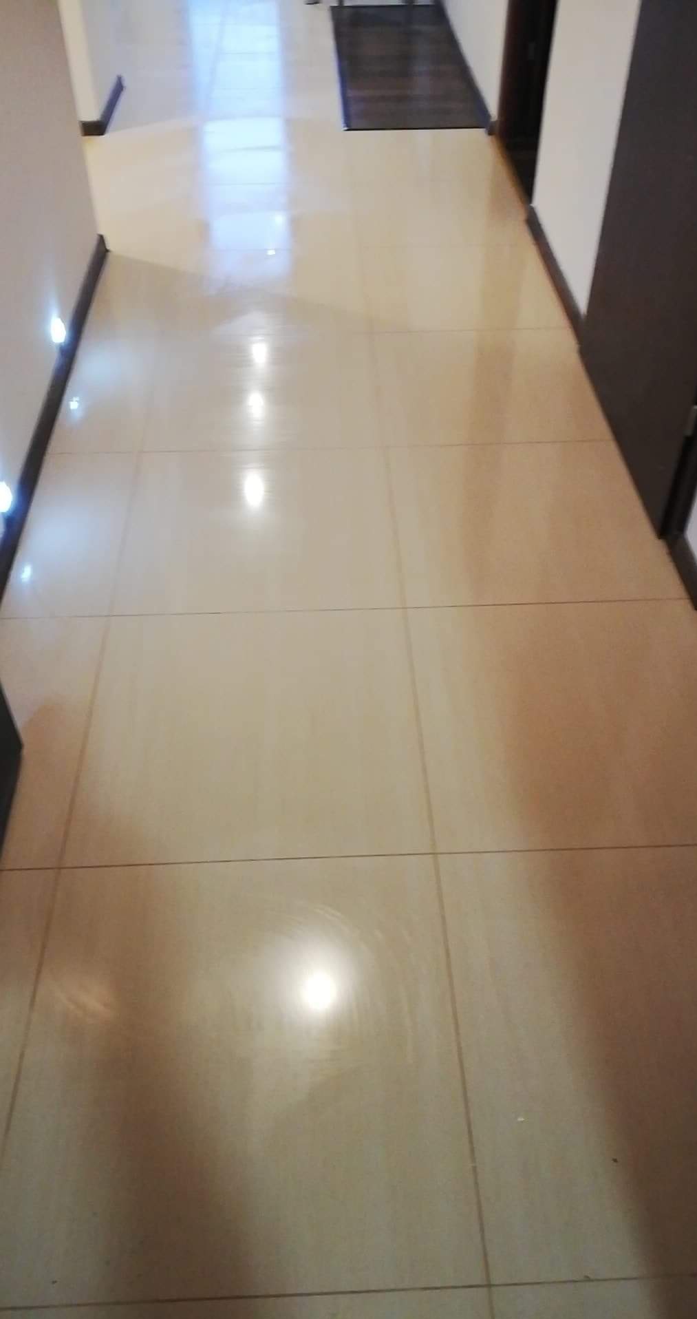 Beżowe, błyszczące płytki podłogowe w korytarzu, z widocznymi listwami przypodłogowymi i oświetleniem punktowym przy ścianie. Perspektywa z góry.