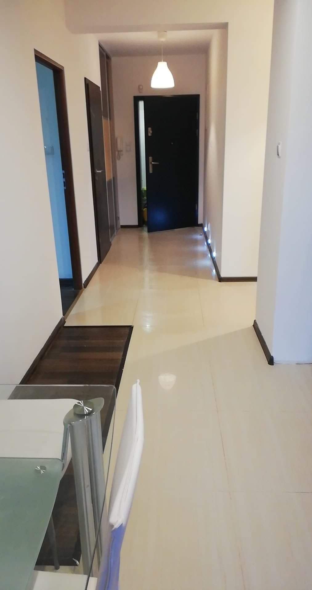 Jasny korytarz z beżowymi płytkami podłogowymi i białymi ścianami. Widoczne drzwi wejściowe i oświetlenie podłogowe. Fragment szklanego stołu w rogu kadru.