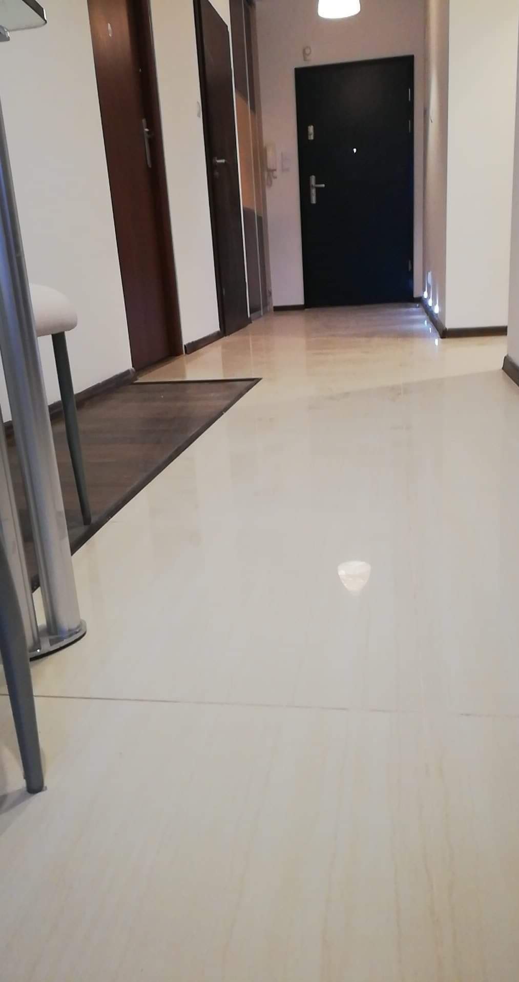 Błyszcząca, kremowa podłoga z płytek w korytarzu mieszkania. Widoczne drzwi wejściowe i wewnętrzne, fragment krzesła. Minimalistyczny design, jasne wnętrze.
