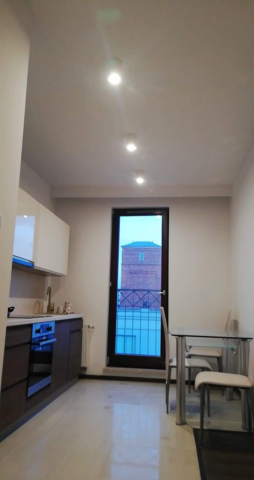 Jasna kuchnia z ciemnymi szafkami i widokiem na ceglaną wieżę przez balkon. Minimalistyczny stół z krzesłami. Podłoga z połyskiem odbija światło.