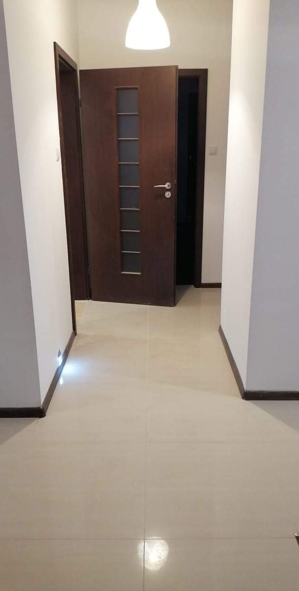 Jasne płytki podłogowe w korytarzu z dwiema drewnianymi ościeżnicami i drzwiami z przeszkleniami. Białe ściany i sufit z lampą.