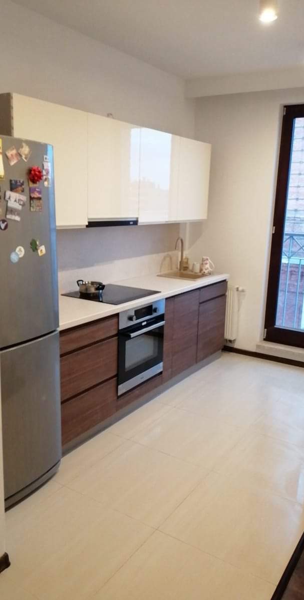 Nowoczesna kuchnia z brązowymi szafkami dolnymi i białymi górnymi, jasnymi płytkami podłogowymi i srebrną lodówką z magnesami. Widok na okno z kratą.