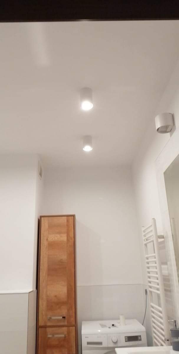 Jasna łazienka po remoncie z drewnianą szafką, pralką i oświetleniem punktowym. Widok na sufit i fragment lustra. Minimalistyczny design.