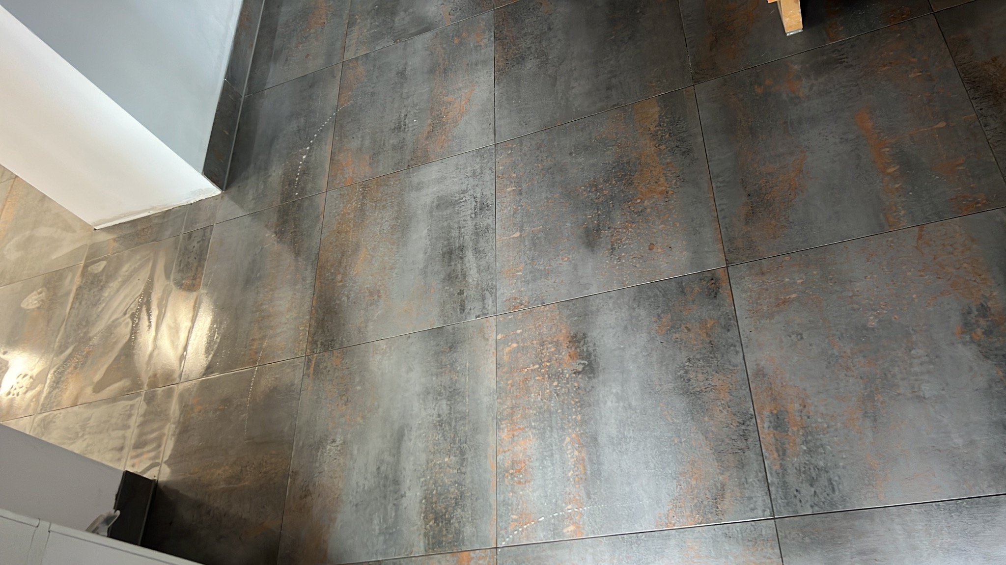 Ułożone, matowe, szare płytki podłogowe z rdzawymi akcentami, widoczne z góry, w rogu fragment białej ściany. Minimalistyczny design, nowoczesne wykończenie.