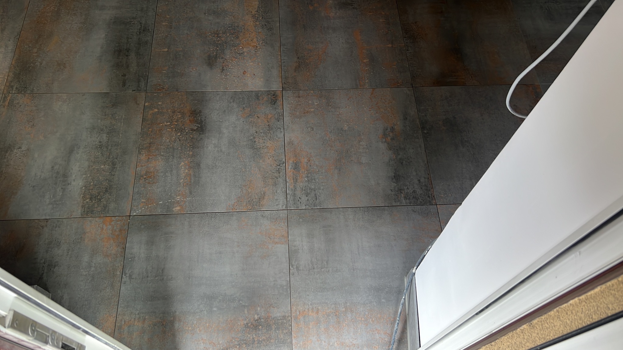 Szare, kwadratowe płytki podłogowe z rdzawymi akcentami, ułożone w łazience. Fragment białej kabiny prysznicowej w prawym górnym rogu kadru.
