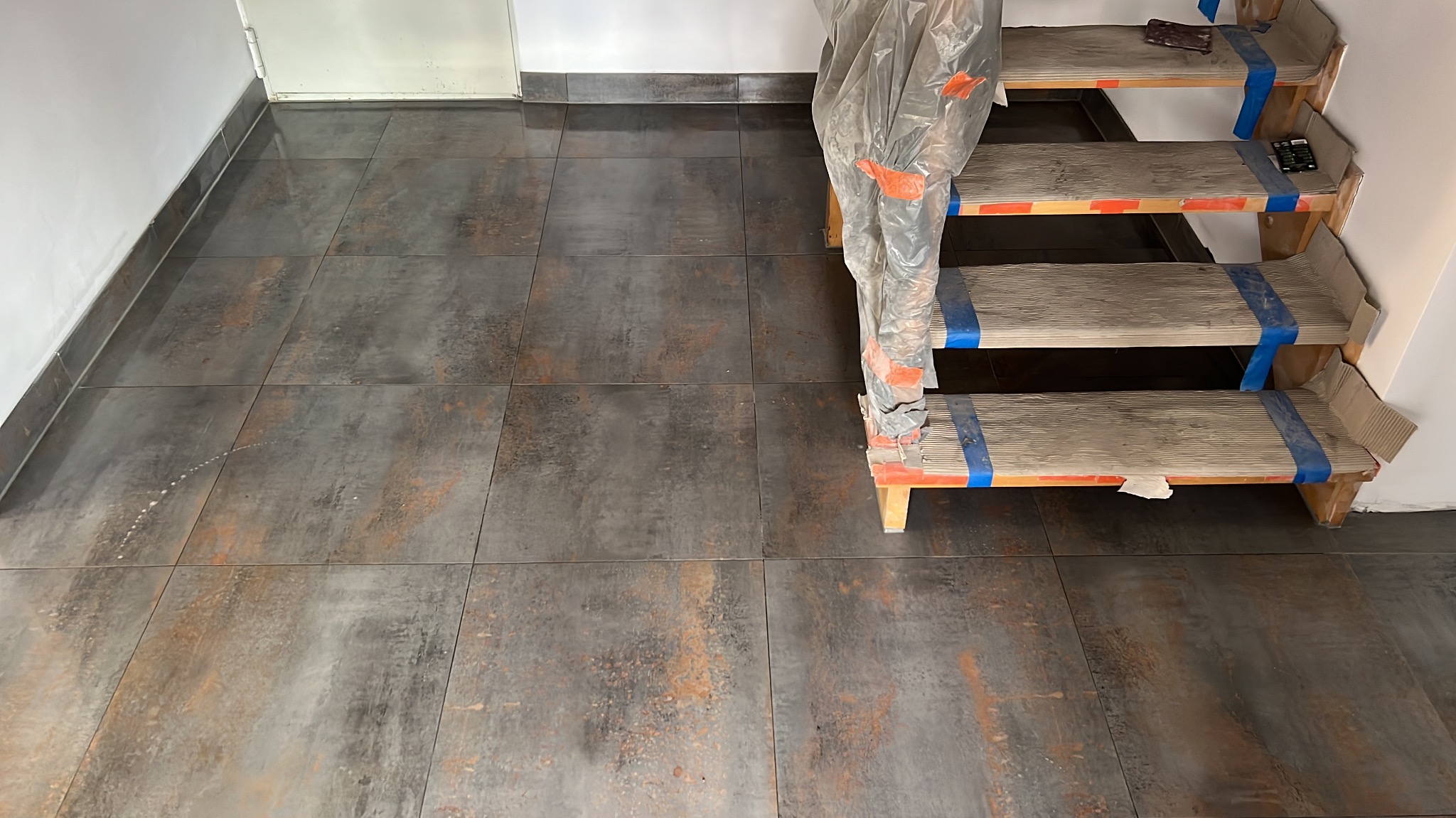Ułożona, ciemna, matowa podłoga z płytek imitujących metal, z widocznymi schodami zabezpieczonymi folią i taśmą. Minimalistyczny design, surowy styl.