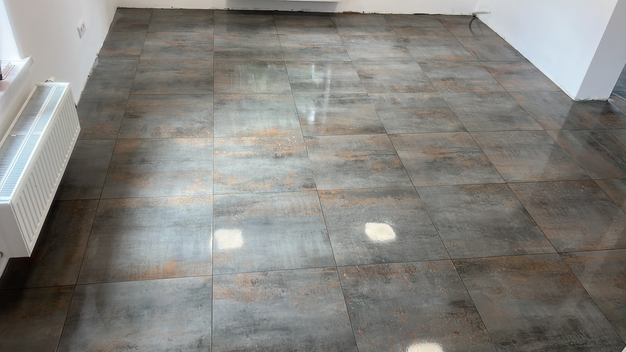 Ułożone, szare płytki podłogowe z efektem rdzy w nowym pomieszczeniu. Widoczny biały grzejnik i fragment ściany. Połyskująca powierzchnia płytek.
