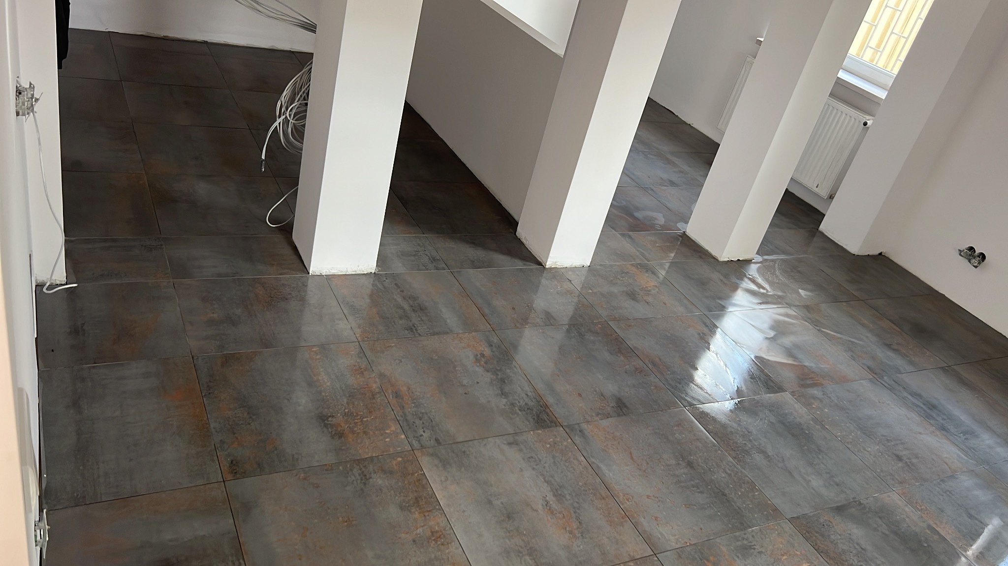 Ułożone, ciemne płytki podłogowe z efektem rdzy w jasnym wnętrzu z białymi kolumnami. Widoczne kable i gniazdka elektryczne.