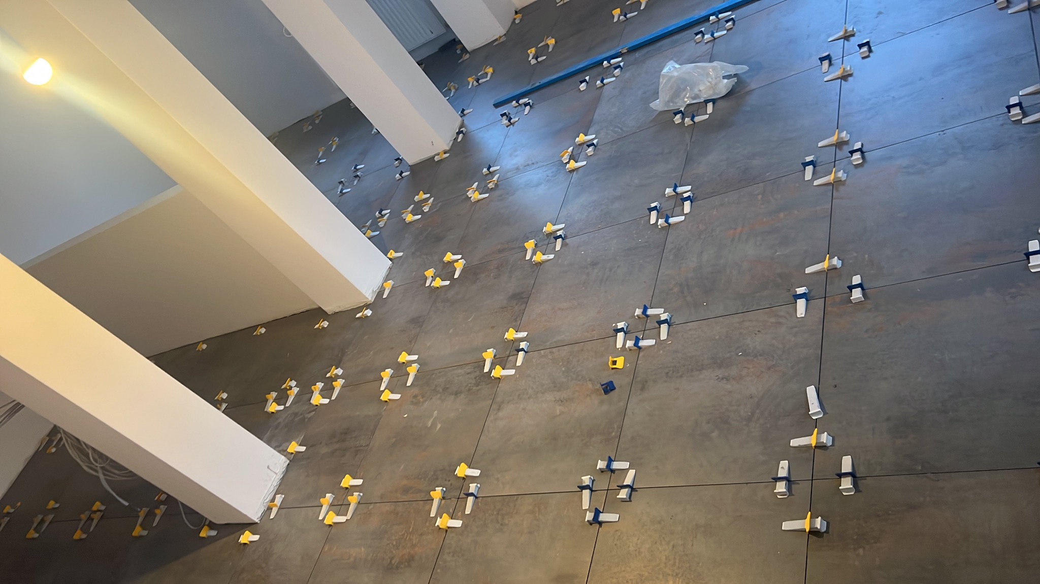 Układanie płytek wielkoformatowych na podłodze z użyciem systemu poziomowania. Widoczne krzyżyki i kliny, szare płytki i białe filary w tle.