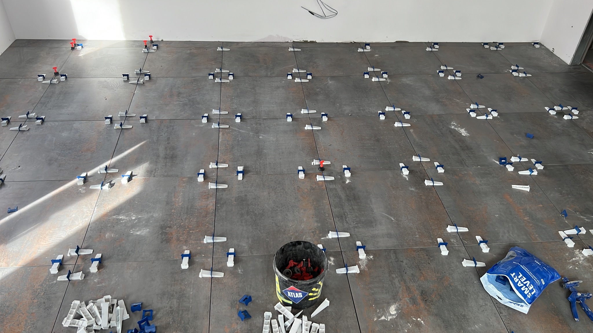 Układanie płytek podłogowych z użyciem systemu poziomowania. Widoczny kubeł z klinami i szczypce do dociskania. Płytki w odcieniach szarości z rdzawym wzorem.