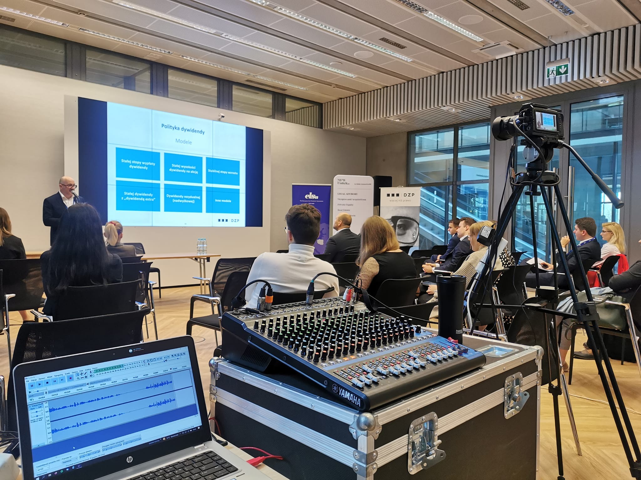 Konferencja z prezentacją na ekranie, nagrywana kamerą na statywie, z mikserem Yamaha i laptopem z oprogramowaniem do edycji dźwięku na pierwszym planie.