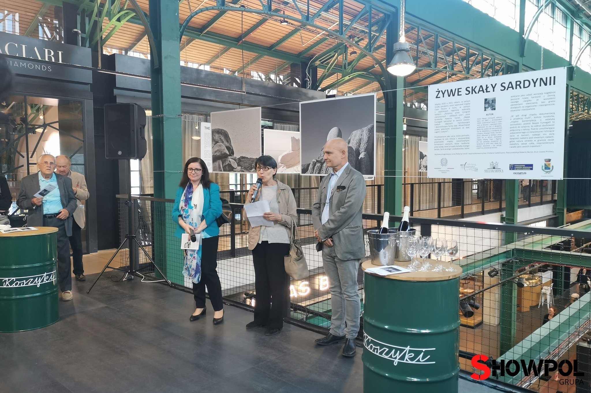 Prezentacja wydarzenia w hali Koszyki: trzy osoby na scenie z mikrofonem, butelki wina w wiaderkach z lodem na beczkach z logo Koszyki, baner z napisem 'Żywe Skały Sardynii' w tle.