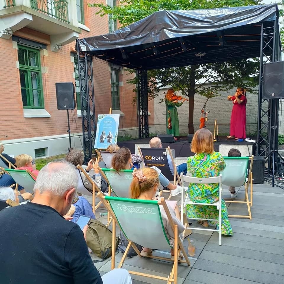 Plenerowy koncert skrzypcowy: dwie skrzypaczki w długich sukniach grają na scenie pod czarnym zadaszeniem, publiczność siedzi na leżakach, z boku widoczny plakat i nagłośnienie.