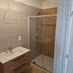 Nowoczesna łazienka z kabiną prysznicową, umywalką z szafką z drewnopodobnym frontem i odsłoniętym przewodem elektrycznym wystającym ze ściany pokrytej szarymi płytkami.