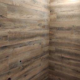 AM usługi wykończeniowe - Wykończona łazienka z drewnopodobnymi płytkami ceramicznymi na ścianach i podłodze, widoczny odpływ liniowy i otwór na instalację elektryczną.