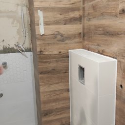 AM usługi wykończeniowe - Wnętrze łazienki w trakcie remontu: ściany wyłożone płytkami imitującymi drewno, zabudowa geberit z otwartym otworem rewizyjnym, fragment ściany z białymi heksagonalnymi płytkami, wzorzysta podłoga...