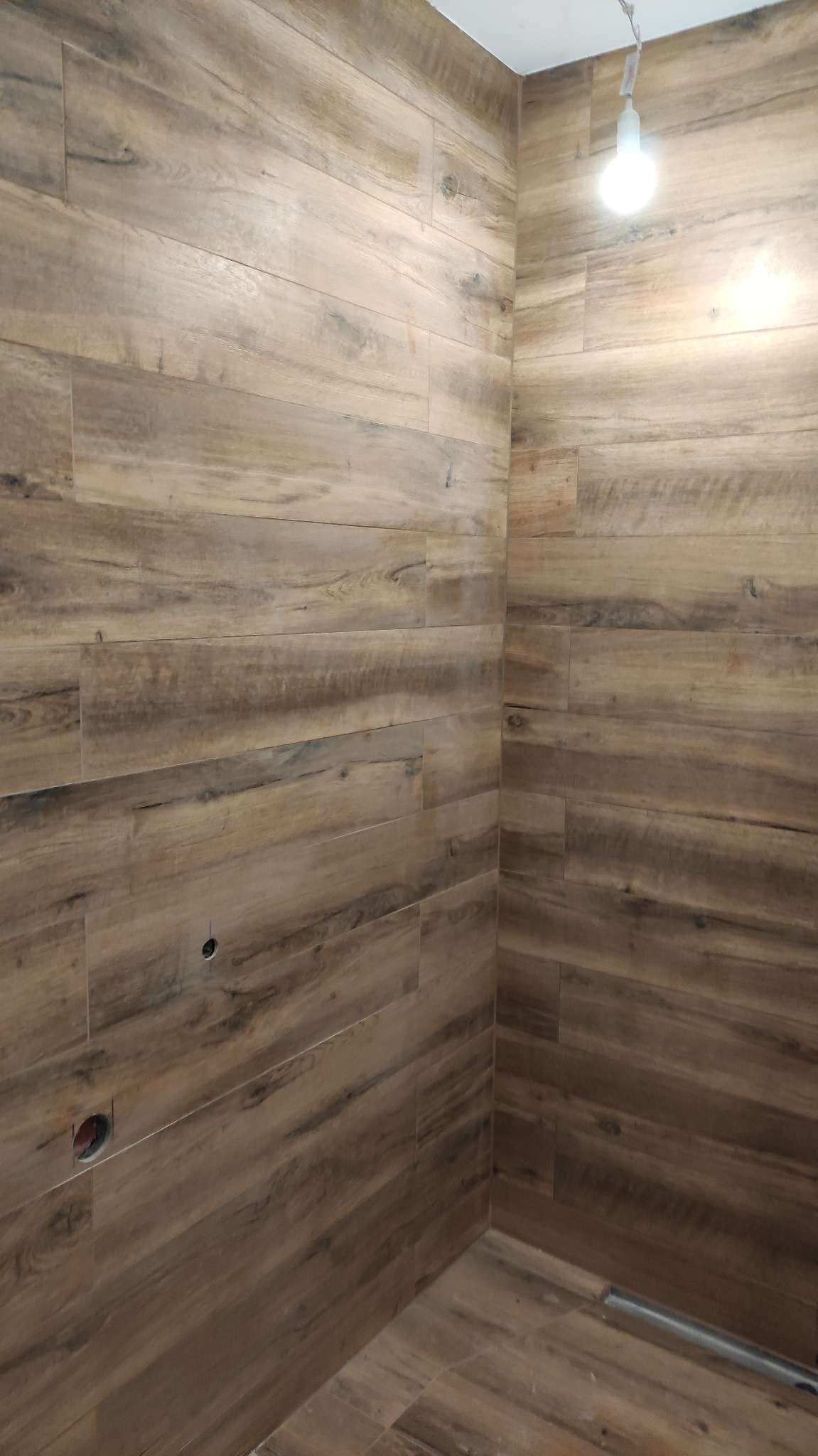 Wykończona łazienka z drewnopodobnymi płytkami ceramicznymi na ścianach i podłodze, widoczny odpływ liniowy i otwór na instalację elektryczną.