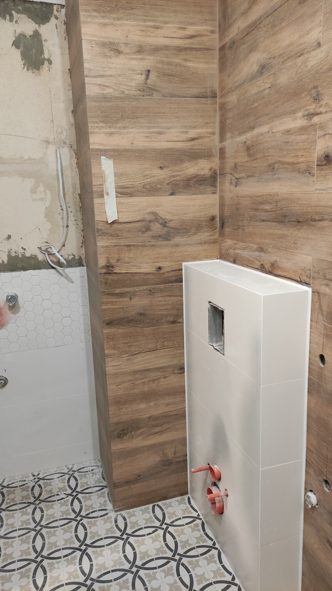 Wnętrze łazienki w trakcie remontu: ściany wyłożone płytkami imitującymi drewno, zabudowa geberit z otwartym otworem rewizyjnym, fragment ściany z białymi heksagonalnymi płytkami, wzorzysta podłoga...