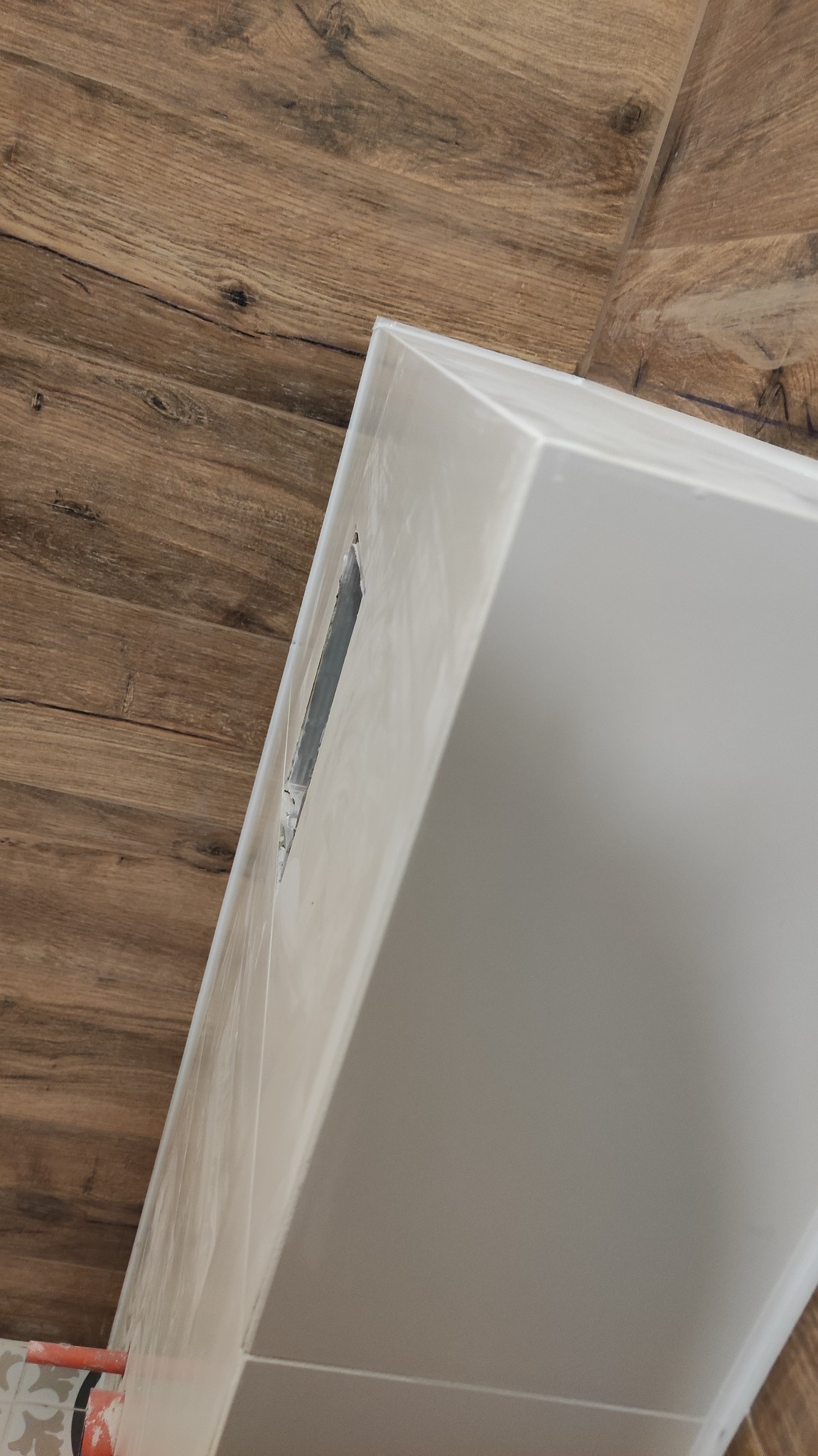 Montaż obudowy geberitu z widocznym otworem rewizyjnym, zabezpieczona folią ochronną, na tle drewnopodobnych paneli ściennych.