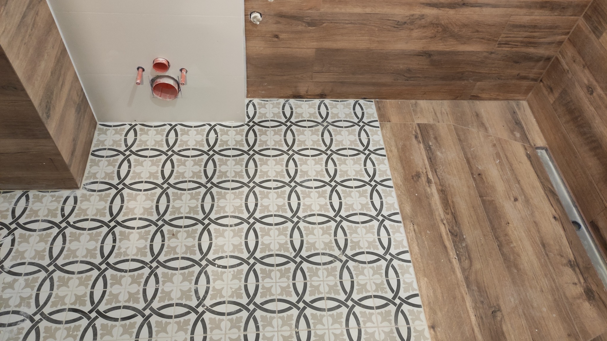 Fragment łazienki w trakcie remontu: wzorzyste płytki podłogowe w odcieniach szarości i beżu, imitacja drewna na ścianie i podłodze, widoczne rury odpływowe w ścianie.