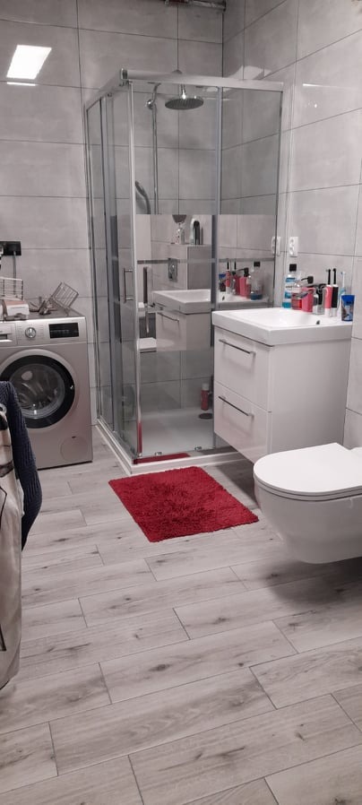 Nowoczesna łazienka z szarymi płytkami imitującymi drewno, kabiną prysznicową typu walk-in, białą umywalką z szafką, wiszącą toaletą i czerwoną matą, pralka z boku.