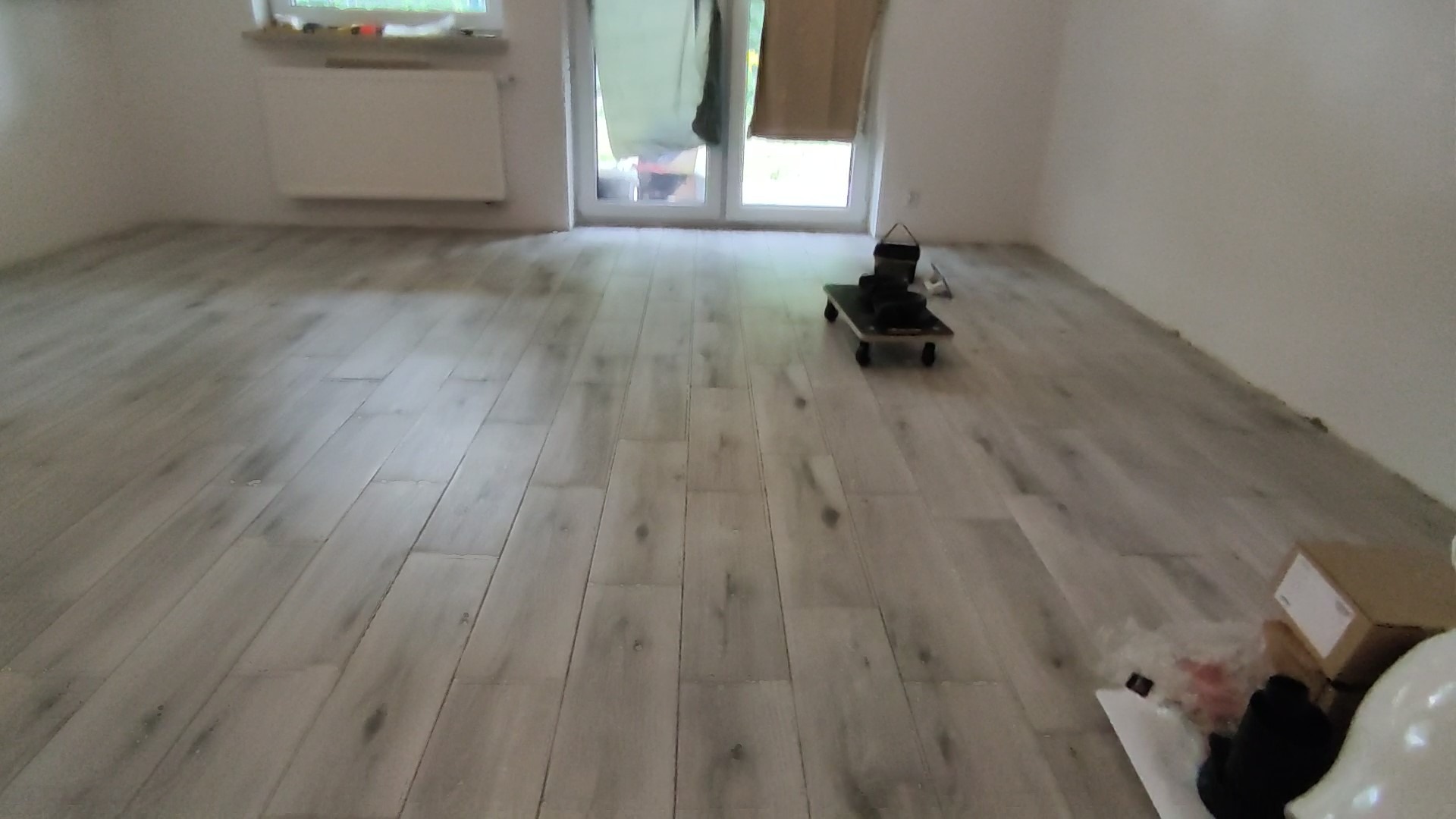 Świeżo ułożona, jasnoszara podłoga z paneli imitujących drewno, widoczne narzędzia glazurnicze na wózku i kartony z materiałami wykończeniowymi przy ścianie.