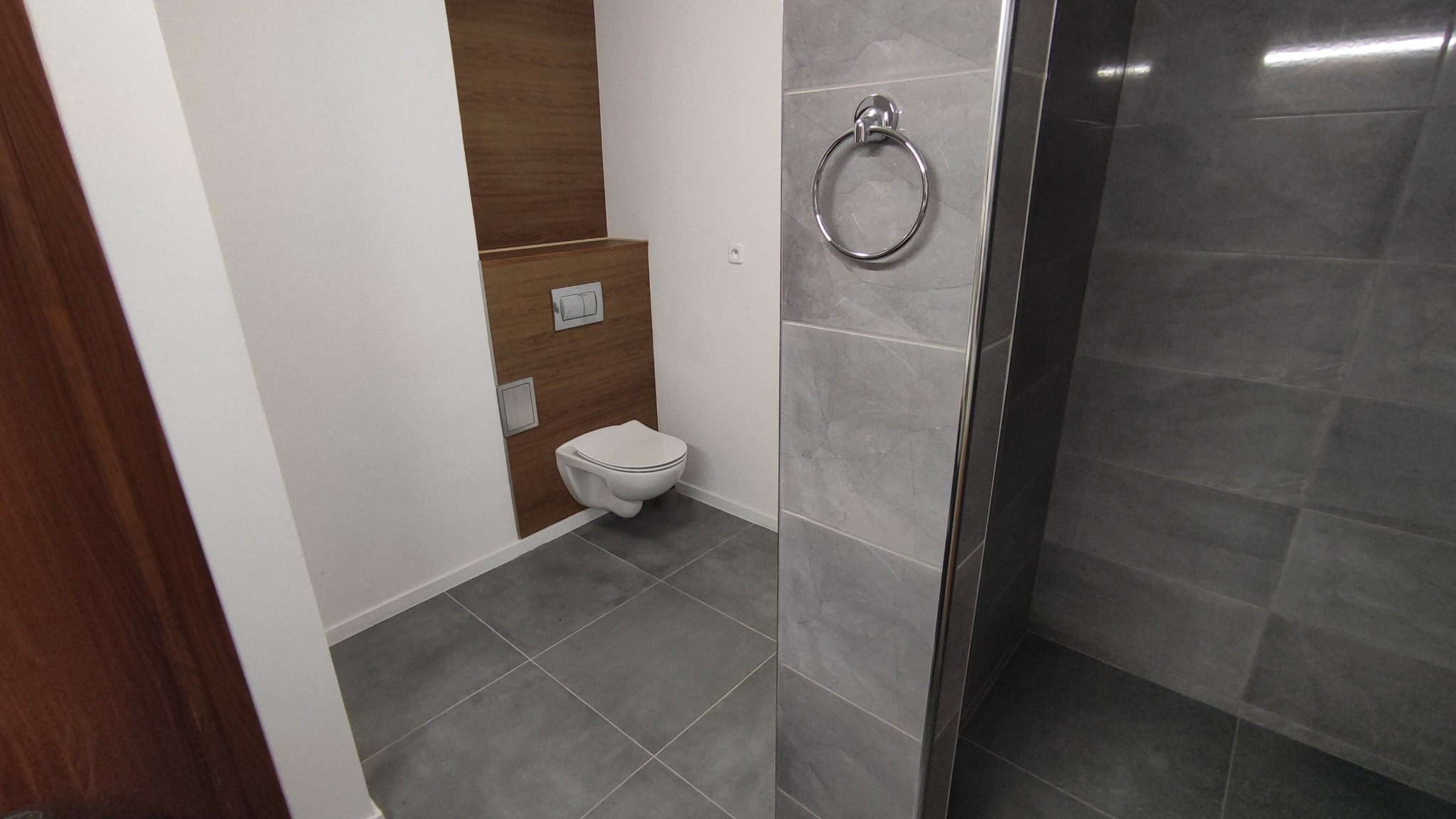 Nowoczesna łazienka z wiszącą toaletą na tle drewnianej ściany, szare płytki podłogowe i ścienne w kabinie prysznicowej, chromowany wieszak na ręcznik.