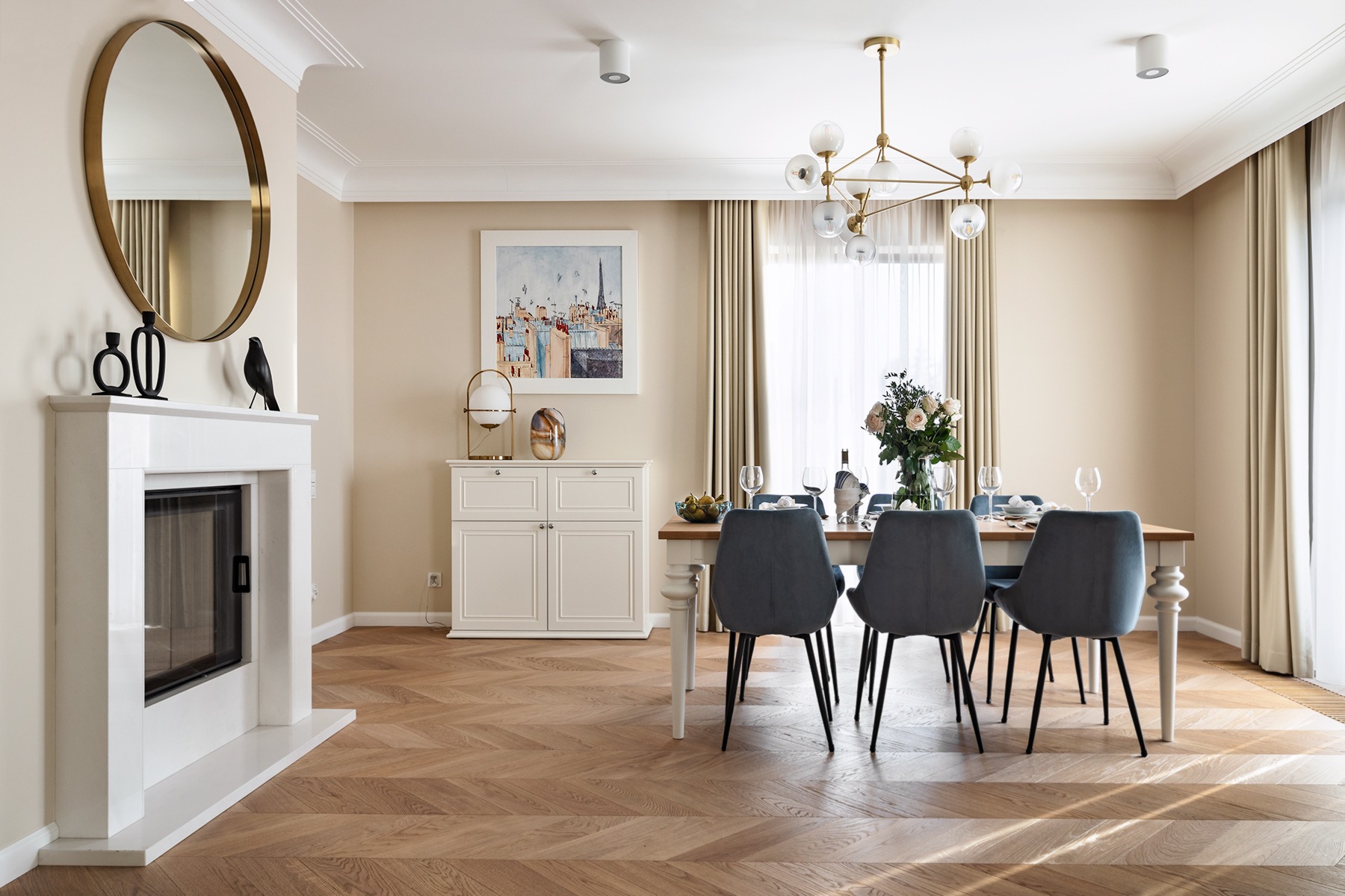 Jasne wnętrze jadalni z parkietem w jodełkę, białym kominkiem z okrągłym lustrem, kredensem, drewnianym stołem z szarymi krzesłami i bukietem kwiatów.