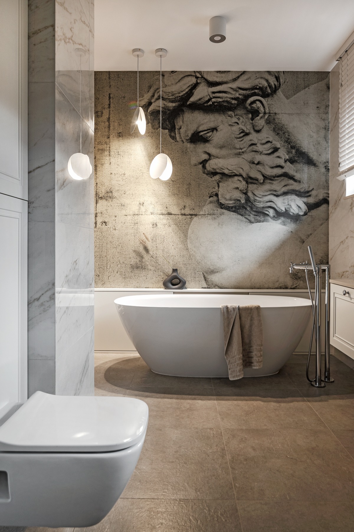 Nowoczesna łazienka z wolnostojącą wanną, marmurowymi płytkami, ceramiczną toaletą i tapetą z motywem rzeźby, oświetlona wiszącymi lampami.