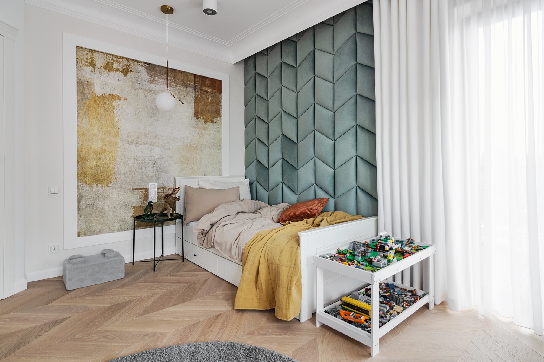 Jasny pokój dziecięcy z białym łóżkiem, geometryczną tapicerką i stolikiem z klockami Lego, oświetlony wiszącą lampą i naturalnym światłem.