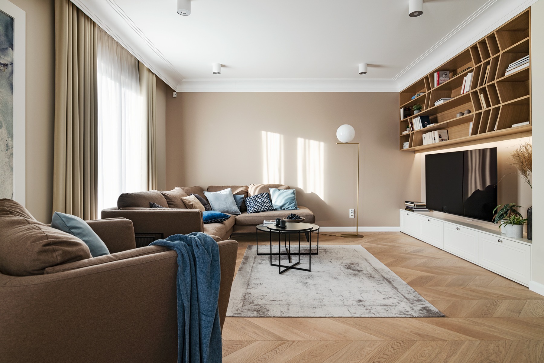 Jasne wnętrze salonu z parkietem w jodełkę, brązową sofą narożną z poduszkami, stolikiem kawowym, regałem z książkami nad telewizorem i lampą podłogową.