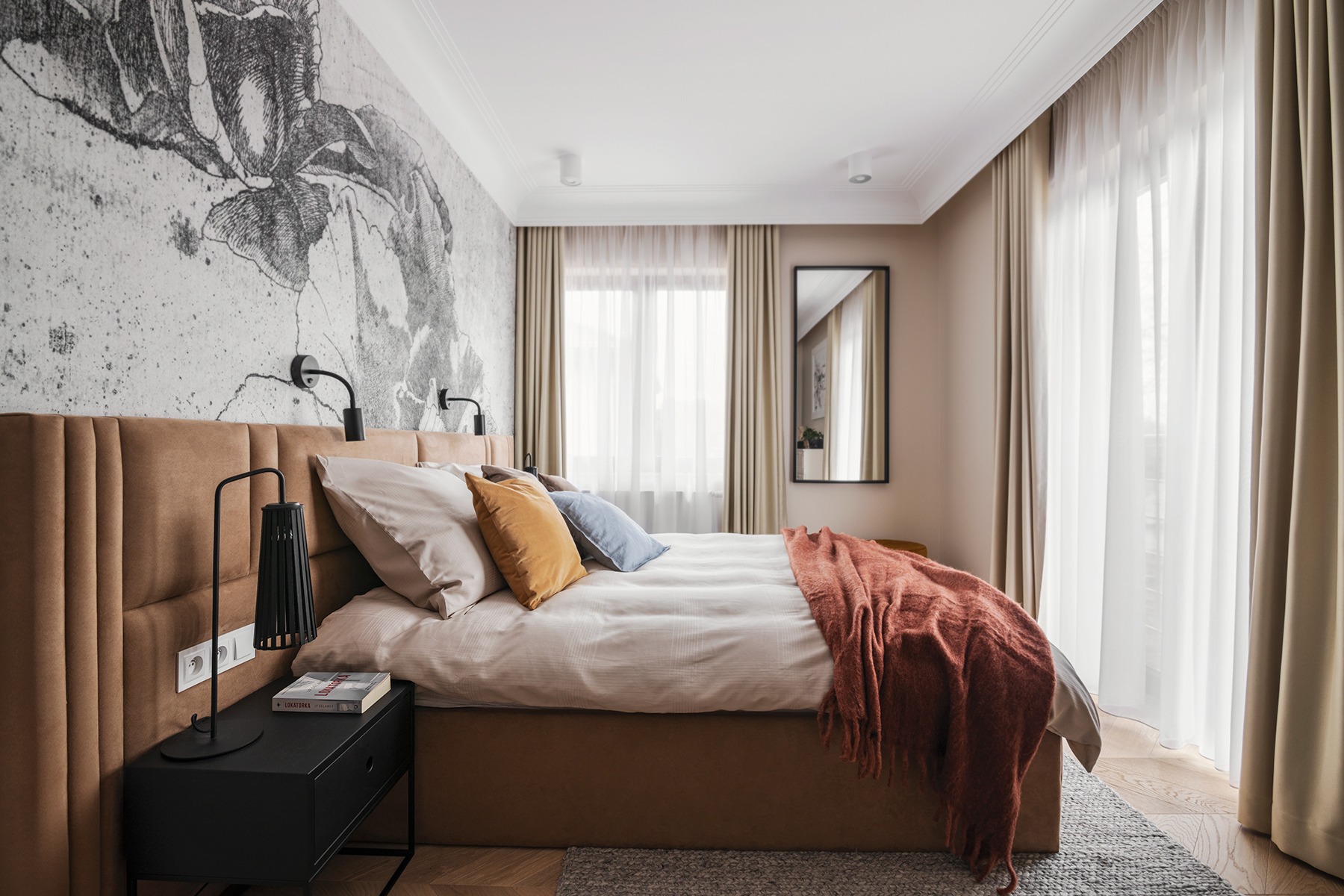 Sypialnia z tapetą w motywy roślinne, brązowym tapicerowanym łóżkiem, czarną szafką nocną z lampką i książką, oraz pomarańczowym pledem na łóżku