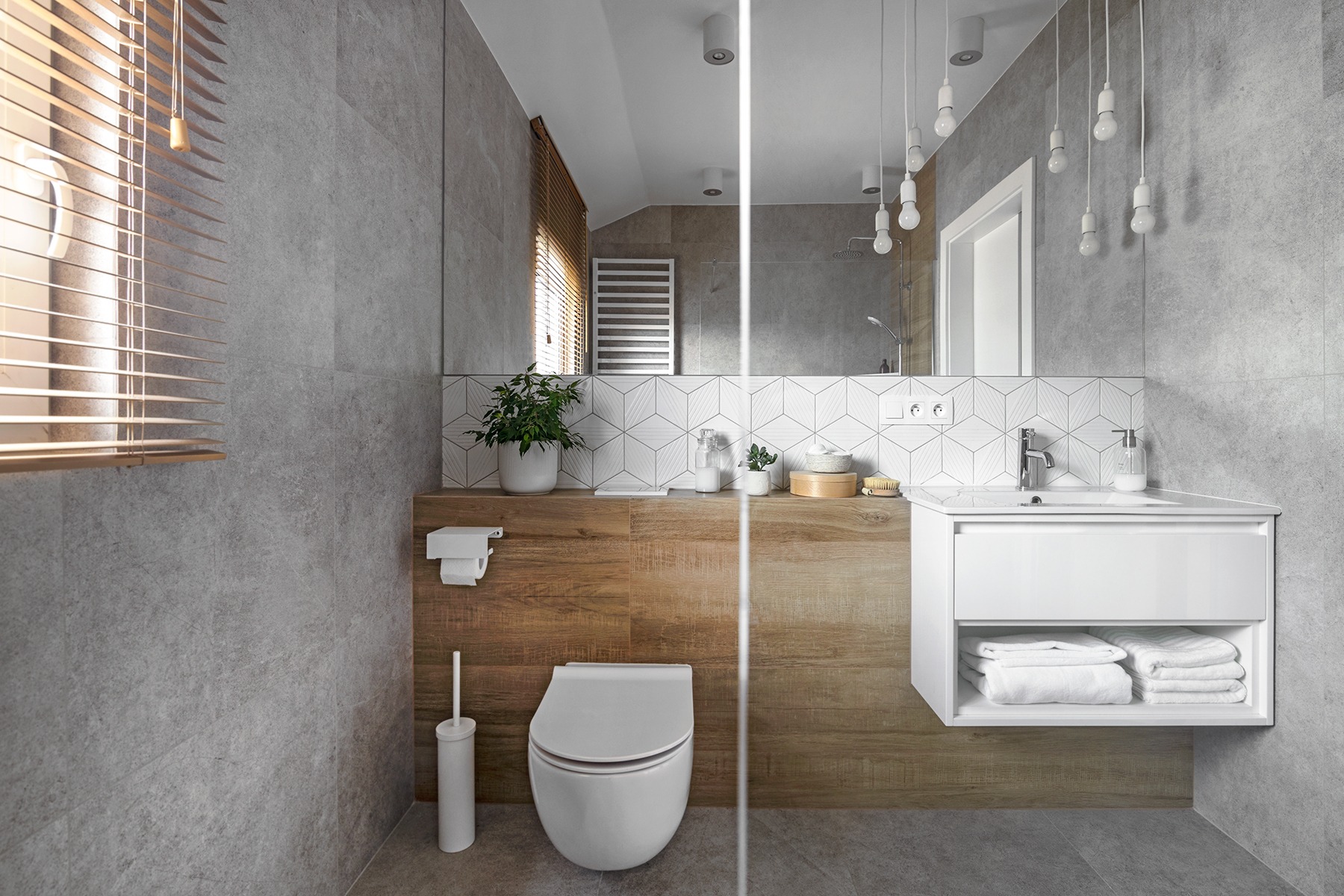 Nowoczesna łazienka z szarymi płytkami, drewnianymi akcentami i białymi elementami wyposażenia, w tym wisząca szafka z umywalką i toaleta, uzupełniona oświetleniem w postaci wiszących żarówek...