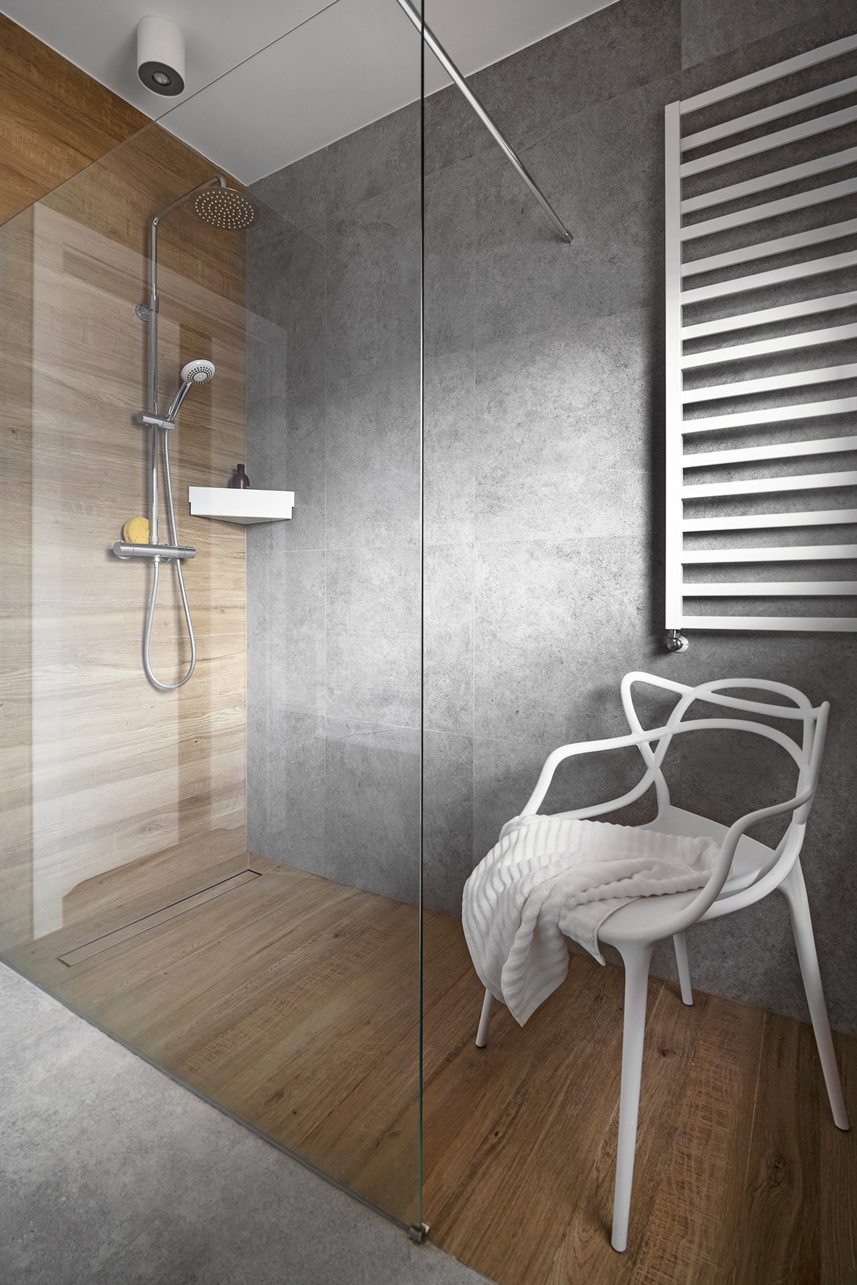 Nowoczesna kabina prysznicowa z drewnopodobnymi płytkami, szara ściana, biały grzejnik drabinkowy i designerskie krzesło z ręcznikiem.