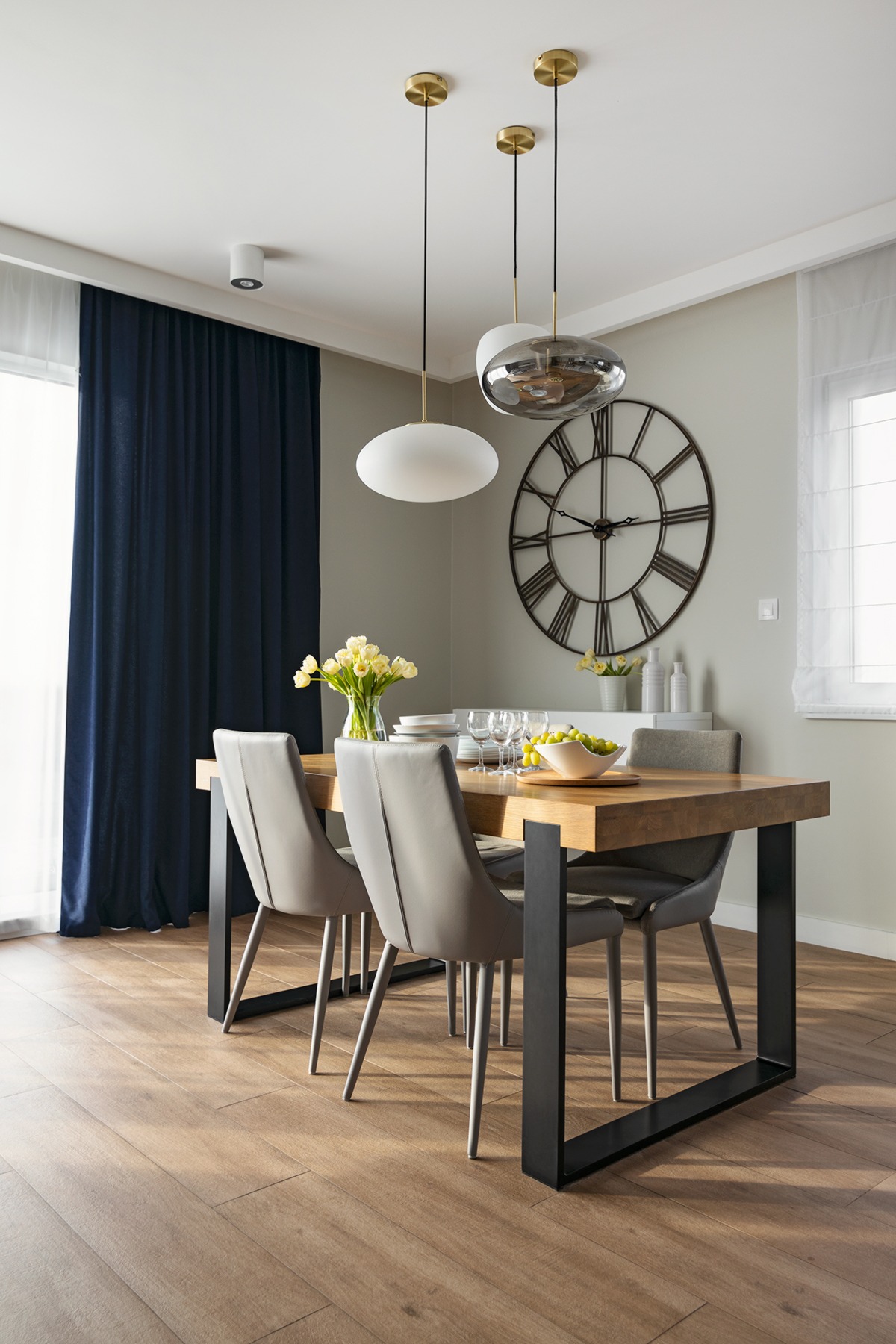 Jadalnia z drewnianym stołem na czarnych nogach, szarymi krzesłami, trzema nowoczesnymi lampami wiszącymi i dekoracyjnym zegarem na ścianie.