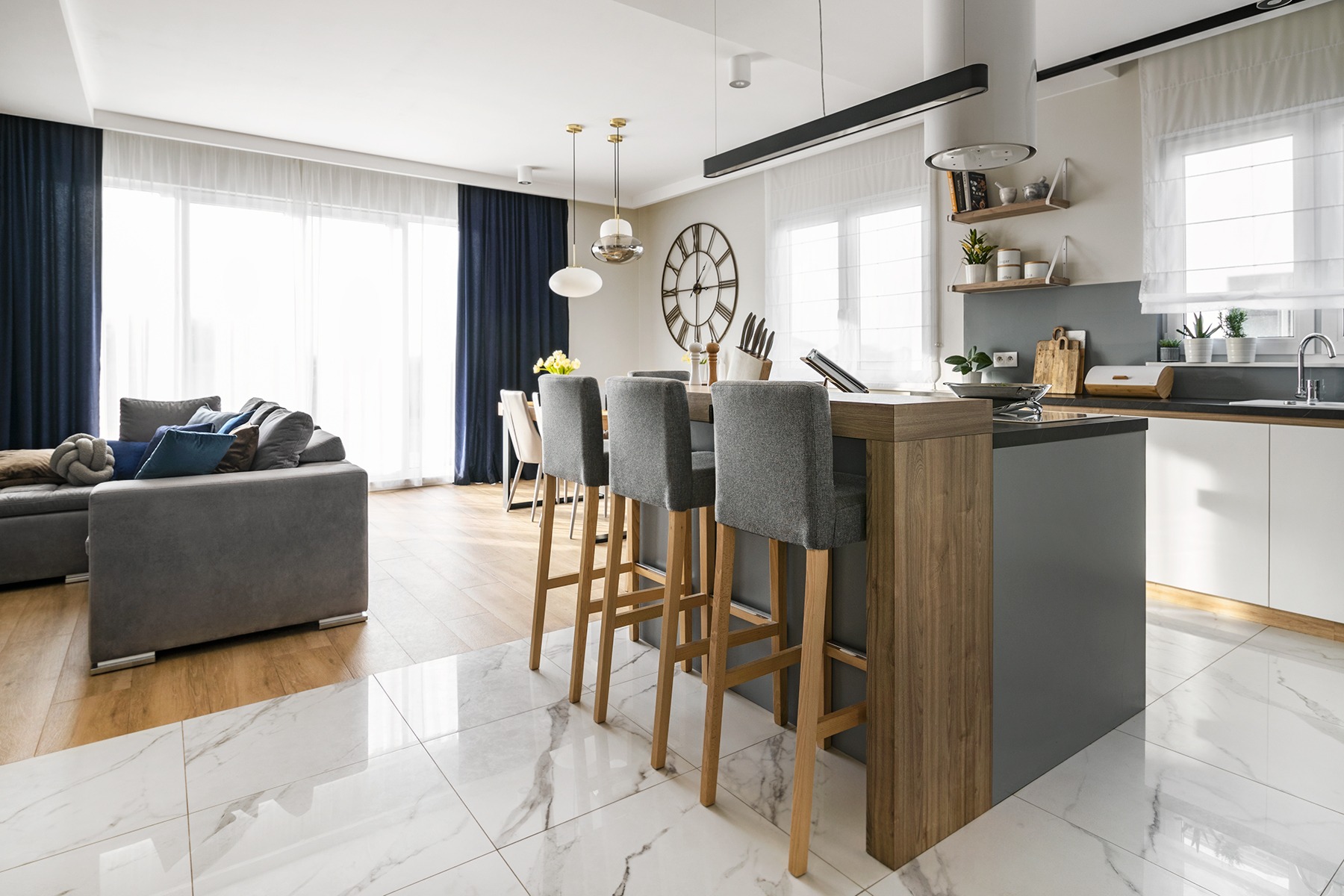 Nowoczesna kuchnia z wyspą i hokerami w jasnym salonie w Łodzi, widoczny fragment sofy i okno z granatowymi zasłonami.