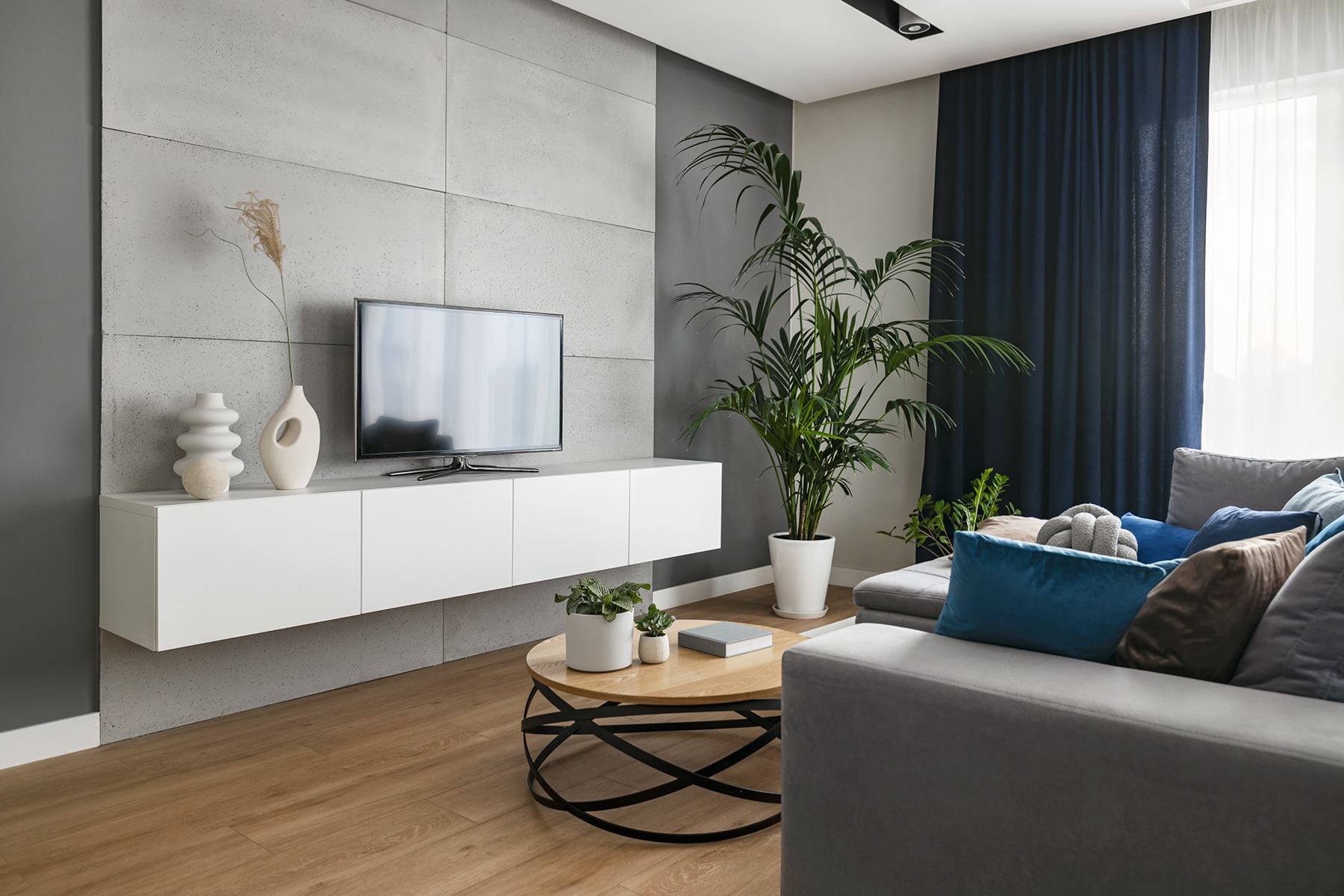 Nowoczesny salon z betonową ścianą, białym regałem pod telewizorem, okrągłym stolikiem kawowym i szarą sofą z niebieskimi poduszkami.