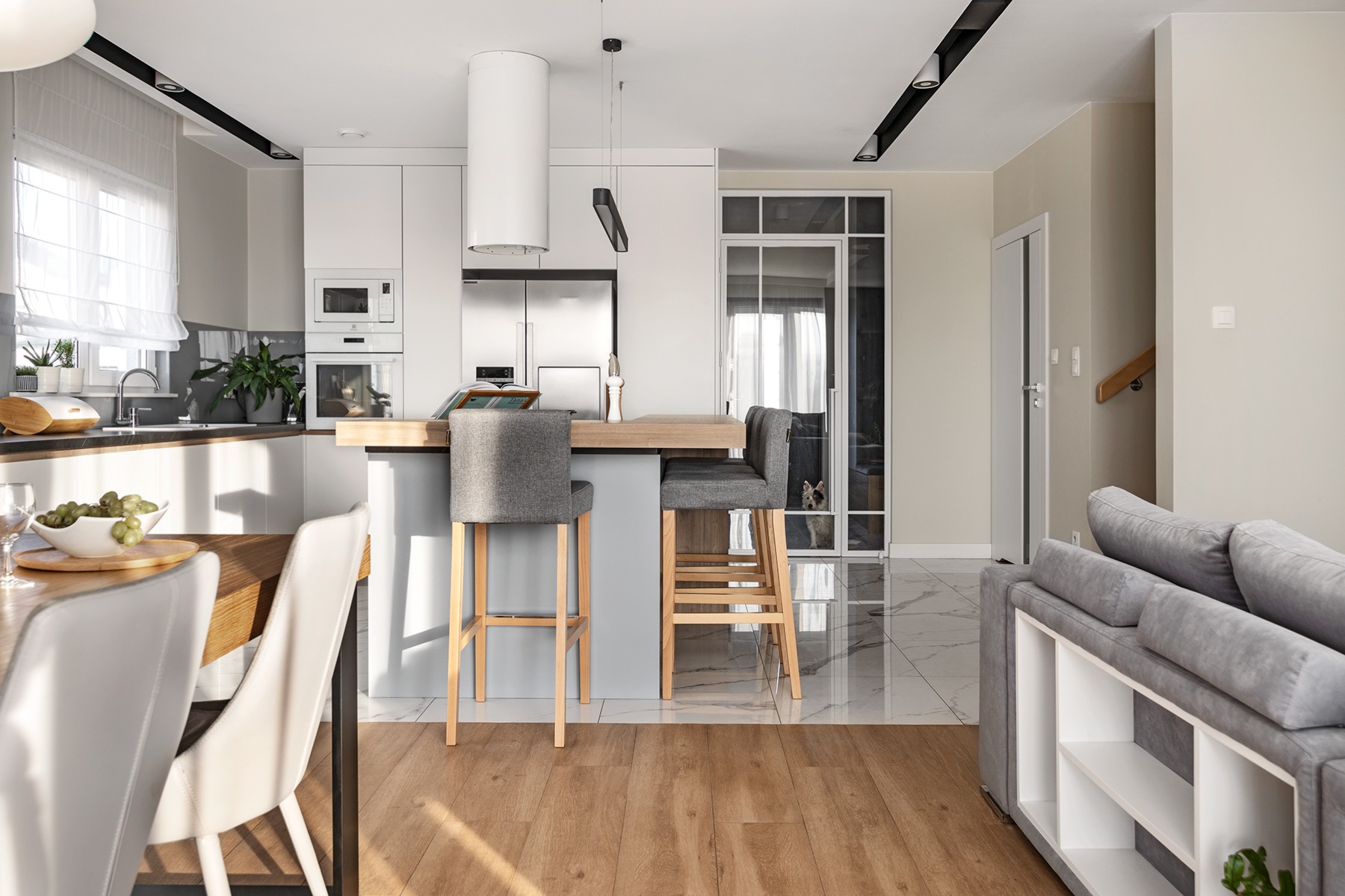 Nowoczesna kuchnia z wyspą i szarymi hokerami, widok na salon z szarą sofą, marmurowa podłoga w przejściu, pies spoglądający przez szklane drzwi.